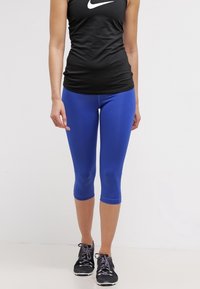 Czarny damski top bez rękawów z białym logo, zestawiony z dopasowanymi niebieskimi legginsami do kolan. Buty czarne z fioletowymi sznurowadłami.