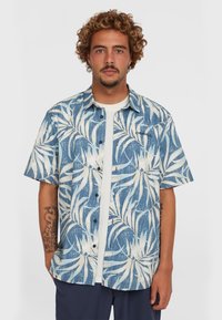 Camisa de manga corta con un patrón de hojas en azul y blanco, cierre de botones en el frente y un ajuste casual, combinada con una camiseta blanca debajo.