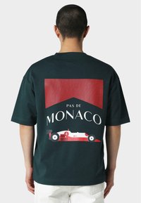 Tume roheline puuvillane T-särk suure graafilise trükiga, millel on punase triibuline taust ja sõiduki disain, mille peal on tekst "PAS DE MONACO".