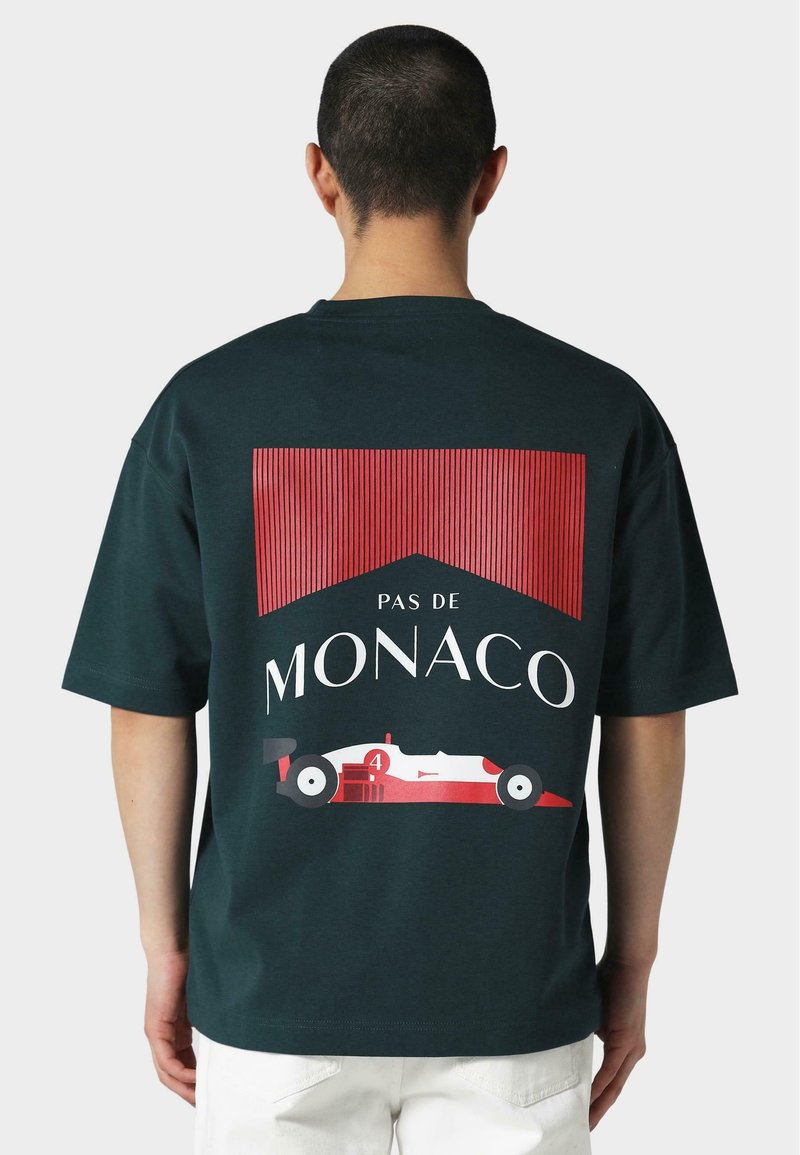 Tume roheline puuvillane T-särk suure graafilise trükiga, millel on punase triibuline taust ja sõiduki disain, mille peal on tekst "PAS DE MONACO".