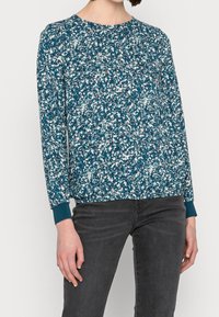 Turkos långärmad topp med blommönster i vitt och rosa, strukturerad tyg och ribbade muddar. I kombination med mörkgråbyxor.