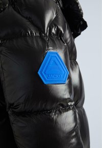 Čierna, lesklá zateplená bunda s hexagonálnym modrým gumeným logom "JACKIT" na hrudi, prešívaný dizajn a hladká textúra.