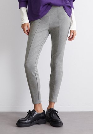 Person wearing slim-fit light gray pants, black lace-up shoes, and a purple top with white long sleeves, standing on gray floor.