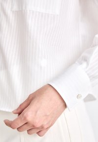 Vit skjorta med ljusa vertikala ränder, en enkel bröstficka och en knappmanschett med en diskret logodetalj. Slät textur.