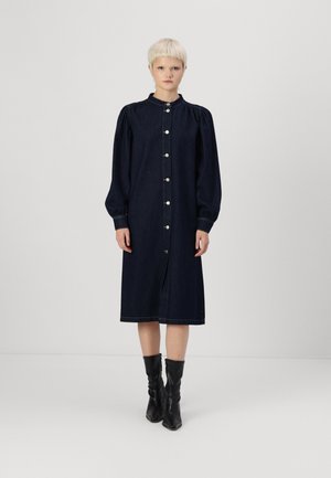 Vestito in denim scuro con maniche lunghe a palloncino, bottoni frontali e colletto, che arriva a metà polpaccio. Abbinato a stivali neri fino al ginocchio.