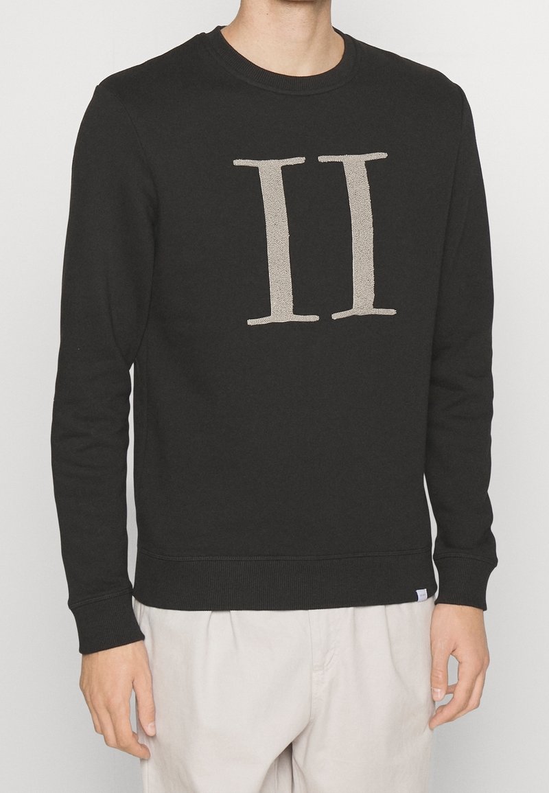 Homme portant un sweat-shirt noir à manches longues avec un grand chiffre romain II beige sur la poitrine, associé à un pantalon clair.