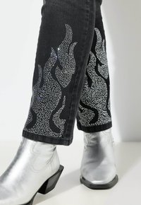 Jeans negros con diseños de llamas de pedrería plateada a lo largo de los lados, combinados con botas metálicas plateadas hasta el tobillo con tacones cuadrados.