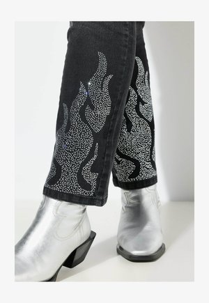Zwarte jeans met zilveren strass vlamontwerpen langs de zijkanten, gecombineerd met metallic zilveren enkellaarsjes met blokhakken.