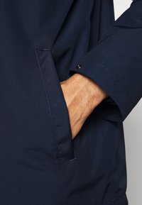 Chaqueta resistente al agua de color azul marino con un acabado texturizado, que presenta un bolsillo lateral y detalles de puños abotonados. La manga está ligeramente enrollada.