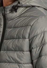 Giacca a vento grigia con design trapuntato, colletto alto, cappuccio con cordoncino e una cerniera nera sulla parte anteriore. La texture appare liscia e imbottita.