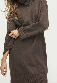 Kvinna justerar ärmen på en brun stickad polotröja med ribbade muddar och nederkant mot en neutral bakgrund.