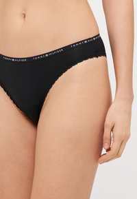 Čierne bikini nohavičky s čipkovým lemom a pásom s potlačou loga. Hladký materiál, priliehavý strih a jemný lesk na textúre tkaniny.