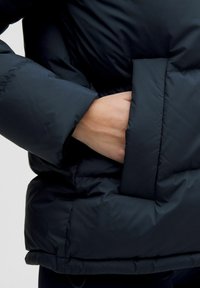 Ruka zasunutá v prednej vrecku čiernej prešívanej bundy, ktorá je oblečená cez tmavé nohavice.