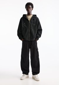 Felpa nera con zip e polsini elasticizzati, abbinata a pantaloni cargo neri larghi con tasche laterali. Indossata con sneakers bianche e una maglia beige.