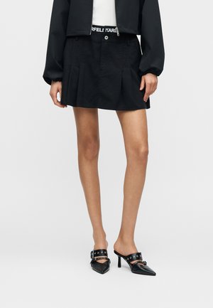 Jupe plissée noire en coton, dotée d'une fermeture par boutons et d'une ceinture avec logo. Associée à des mules noires à bouts pointus avec détails métalliques.