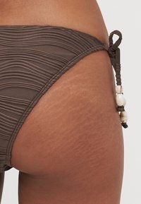 Brown bikini bottom featuring a textured wave pattern, side ties with beads, and a snug fit. The fabric appears smooth and elastic.