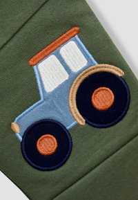 Vihreä kangas, jossa on sininen brodeerattu traktori oransseilla ja ruskeilla yksityiskohdilla, mukaan lukien yksityiskohtaiset renkaat ja ikkunat.