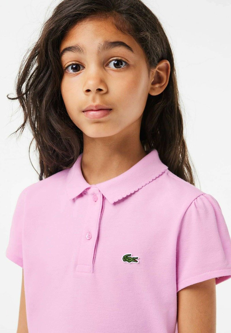 Polo rose clair avec un col festonné, des manches courtes bouffantes et un logo de crocodile brodé vert sur la poitrine gauche.