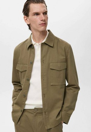 Olive green jacket made of cotton with a collared neckline, button closure, two chest pockets, and long sleeves. Worn over a white top.