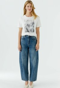 Wit T-shirt met een zwart-wit grafische print, gecombineerd met losse blauwe denim jeans. Eenvoudig ontwerp en een ontspannen pasvorm. Model draagt beige schoenen.