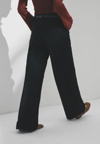 Plati melni bikses, izgatavotas no teksturēta auduma, ar aizmugures kabatām un piegulošu vidukli. Brūnas plakanas kurpes papildina apģērbu.