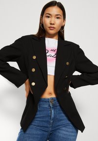 Mustavalkoinen kaksirivinen bleiseri, jossa on kultanapit, rakenteelliset olkapäät ja ylisuuria hihoja, yhdistettynä korkeavyötäröisiin sinisiin farkkuihin.