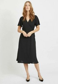 Crna haljina na preklop s kratkim lepršavim rukavima, dužine do sredine lista, izrađena od laganog materijala s vertikalnim rebrastim uzorkom, uparena s crnim štiklama.