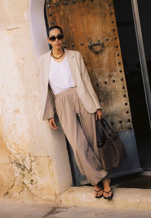 Femeie cu sacou bej și pantaloni largi, purtând ochelari de soare și sandale, sprijinită de un perete texturat lângă o ușă mare de lemn, ținând o geantă maro.