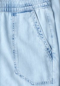 Tissu en denim bleu clair avec une poche d’angle cousue et un détail de ceinture élastique sur un pantalon.