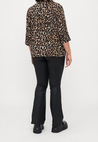 Chemise à imprimé léopard avec des manches trois-quarts, associée à un pantalon évasé noir et des chaussures noires à semelles épaisses. Fond neutre.