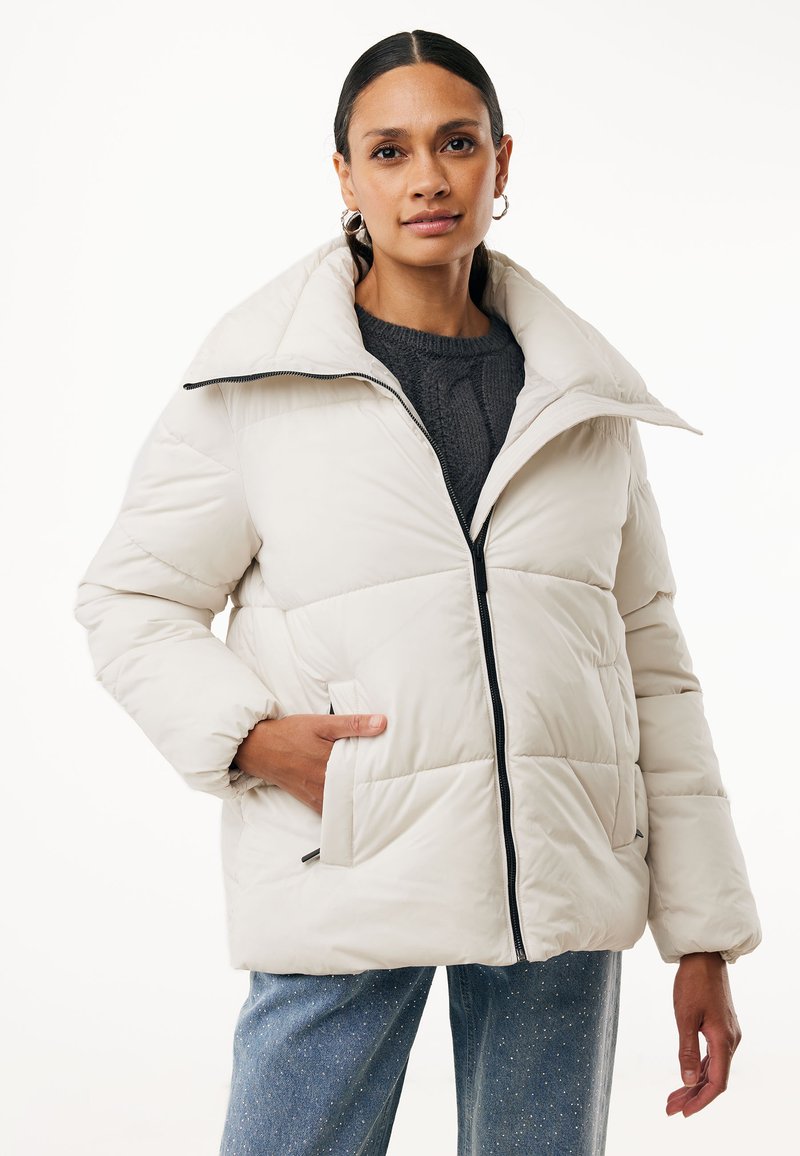 Giacca a vento color beige chiaro con colletto alto, chiusura con zip, tasche frontali e texture trapuntata, indossata sopra un maglione scuro e jeans blu.