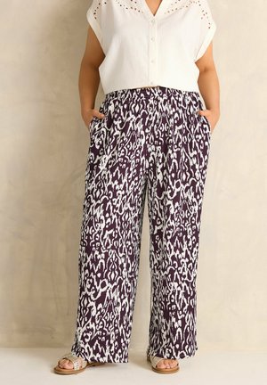 Person iført hvid kortærmet bluse og vidbenet mønstrede bukser med hænderne i lommerne, stående mod en ensfarvet beige væg.