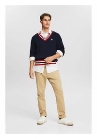 Maglione blu navy a trecce con scollatura a V a righe bianche e rosse, indossato sopra una camicia bianca a maniche lunghe, abbinato a pantaloni beige e sneakers bianche.