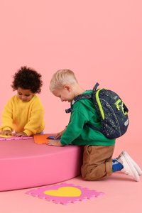 Two children play on a pink, round mat. One wears a yellow knit sweater; the other sports a green hoodie and a patterned navy backpack with neon accents.