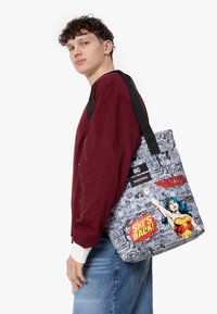 Wonder Woman tygväska med grått serietidningsmönster, med färgglada grafik och text. Svarta bärremmar för enkel transport, strukturerad form.