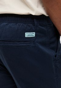 Κοντινό πλάνο ενός ατόμου που φοράει σκούρο μπλε παντελόνι με μια μικρή ανοιχτό μπλε ετικέτα Marc O'Polo Denim πάνω από την πίσω τσέπη.