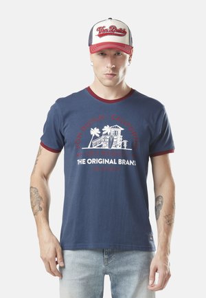 T-shirt en coton bleu marine avec des accents rouges et un imprimé graphique mettant en avant des palmiers. Porté avec un jean bleu clair et une casquette.