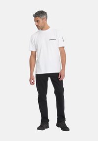 Hombre con camiseta blanca de manga corta Spyder, pantalones negros y zapatos negros, de pie contra un fondo claro y liso.