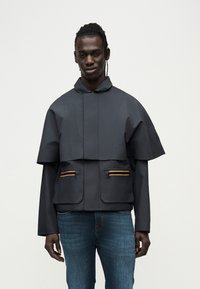 Σκούρο γκρι μπουφάν με κοντό σχέδιο, φαρδιά μανίκια, πορτοκαλί λεπτομέρειες στις τσέπες και ψηλό γιακά. Λείο, ελαφρύ ύφασμα.