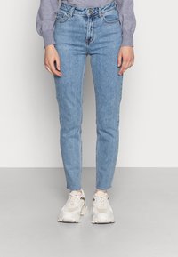 Jeans de tiro alto de color azul claro con un corte ajustado, que cuentan con cinco bolsillos, costuras visibles y una textura ligeramente desgastada. Combinados con zapatillas beige.