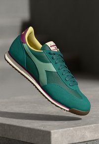 Teal en groene sportshoe met een bovenwerk van mesh, gele binnenvoering, paarse hielaccent en een bruine rubberen zool. Kenmerkt zich door een diagonaal logo ontwerp.