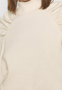 Beige tröja med hög krage, sammetslika puffärmar och slät tygtextur. Enkelt design utan märkbara mönster eller detaljer.