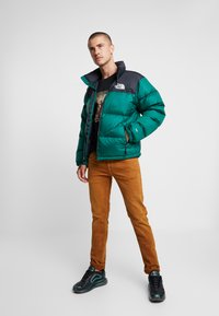 Grøn pufferjakke med sort krave, med lynlås og håndlommer, parret med brune bukser og sorte sneakers.