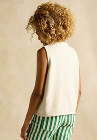 Krémový bezrukávový fleece top s vysokým golierom, v kombinácii so zelenými a bielymi pásikavými nohavicami. Na látke je viditeľná mäkká textúra.