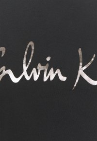 Černá tkanina s metalickým stříbrným kurzívním textem "Calvin Klein." Textura je hladká s jemným leskem. Jednoduchý a minimalistický design.
