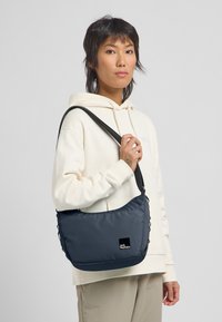 Borsa a spalla blu scuro con superficie testurizzata, tracolla regolabile e patch con logo nero, tenuta da una persona in una felpa color crema.