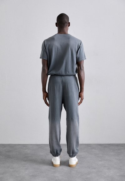 Costum în două piese de culoare gri, realizat dintr-un material moale, compus dintr-un tricou cu croială lejeră și pantaloni cu șnur la brâu, având manșete elastice, asortați cu adidași albi.
