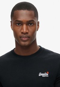 T-shirt nera in cotone a maniche corte con logo "SuperDry" bianco e rosso. Tessuto liscio con vestibilità classica e scollo a costine.