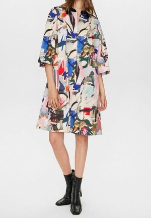 Mujer con vestido hasta la rodilla con estampado abstracto colorido, mangas tres cuartos y botas negras hasta el tobillo, de pie contra un fondo liso.