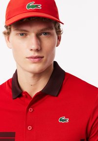 Röd polo t-shirt med en mörkbrun krage, prydd med en liten grön Lacoste-krokodil-logga. Slät textur med en klassisk knappslå.